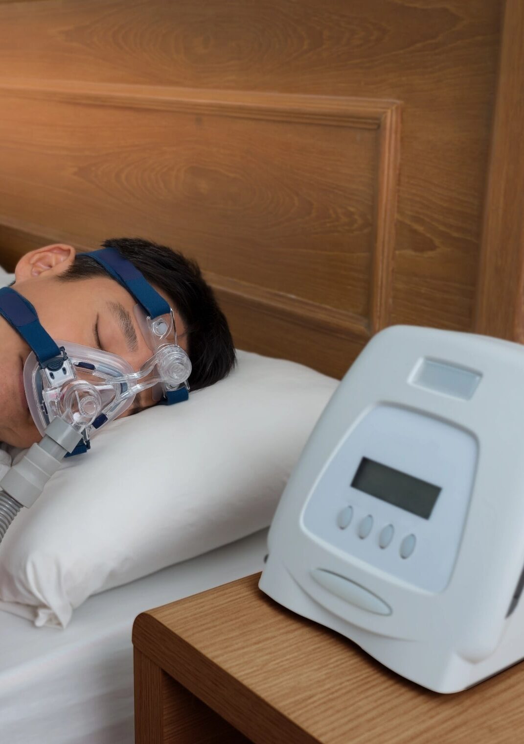 Mężczyzna śpi w masce tlenowej CPAP, obok na stoliku stoi aparat do terapii bezdechu sennego.
