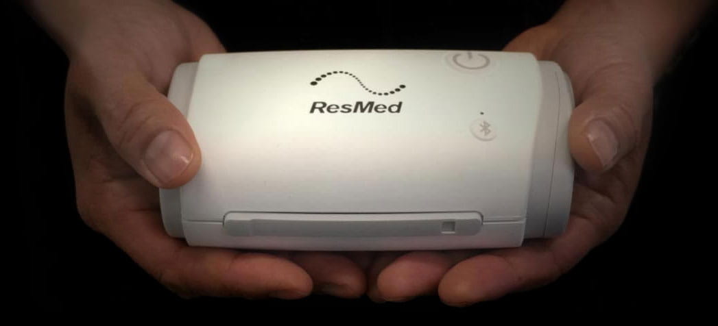 AirMini (1) Dwie dłonie trzymają przenośny aparat CPAP marki ResMed na czarnym tle.