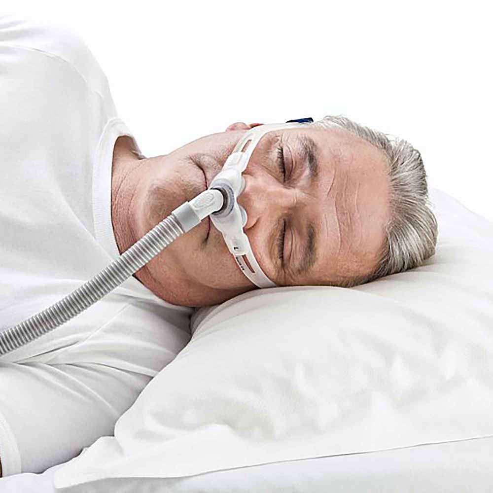 bezdech-nocny-maska-cpap-donosowa-swift-fx-resmed Mężczyzna śpi na boku z maską tlenową CPAP na nosie, leżąc na białej poduszce.
