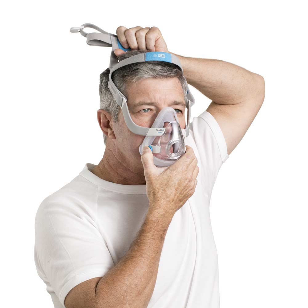 p-EU-63034-ResMed-AirTouch-comfortable-CPAP-mask-03-Medium Mężczyzna zakłada maskę tlenową typu CPAP na twarz.
