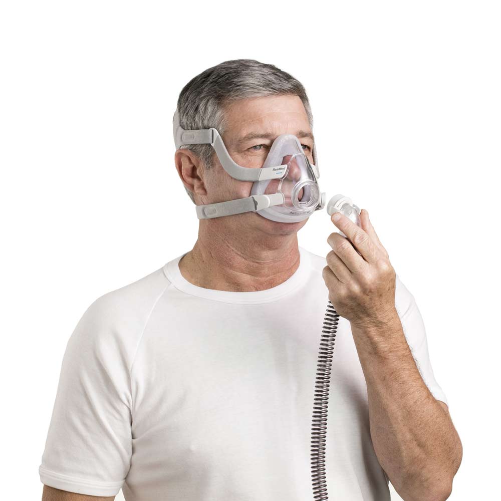p-EU-63034-ResMed-AirTouch-comfortable-CPAP-mask-04-Medium Mężczyzna w białej koszulce zakłada maskę do terapii CPAP na twarz.