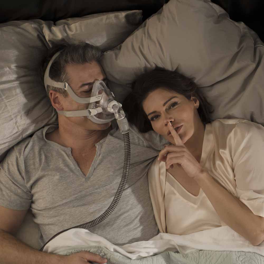 p-EU-63494-ResMed-QuietAir-Quiet-CPAP-Mask-Elbow-03-Medium Mężczyzna śpi w masce CPAP, kobieta obok przykłada palec do ust w geście ciszy.